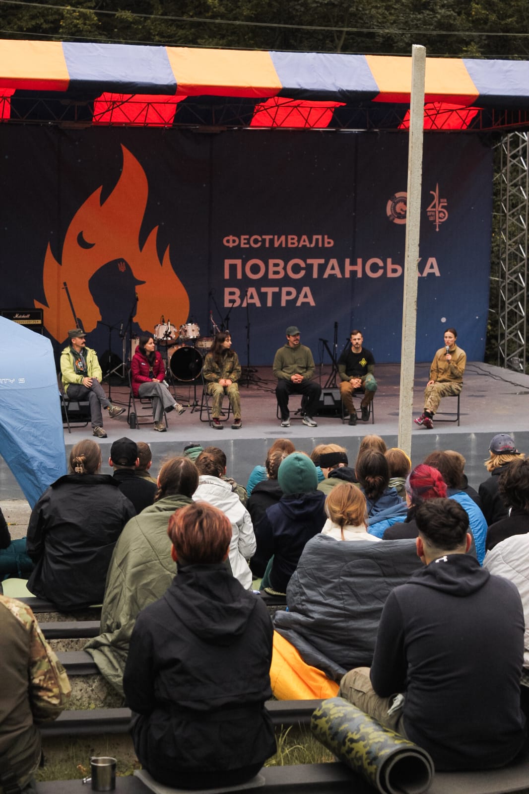 На сцені сидить панель спікерів у військовому та повсякденному одязі, одна жінка говорить у мікрофон. Перед сценою сидить аудиторія молодих людей.