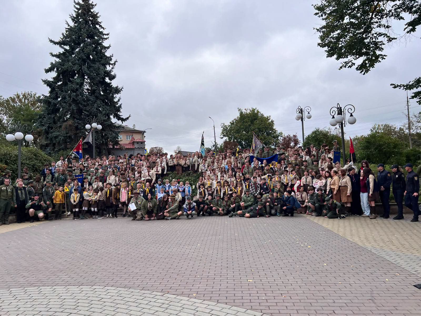 Велика група дітей і дорослих у зеленій пластовій формі з жовтими хустками стоять і сидять на бруківці просто неба. Серед них є прапори, кілька поліцейських праворуч. Позаду дерева, ліхтарі та похмуре небо.