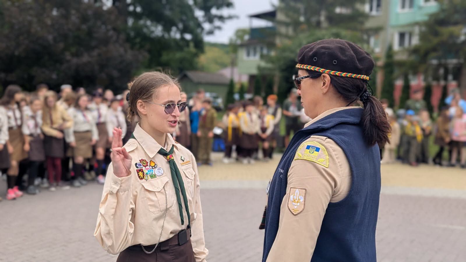 Шмайдей Оксана Василівна, директорка Тернопільського обласного пластового вишкільного центру, стоїть праворуч у пластовій формі. Ліворуч навпроти неї дівчина-пластунка. Обидві підняли руку для пластунського вітання трьома пальцями. На задньому плані група дітей і підлітків у пластовій формі.