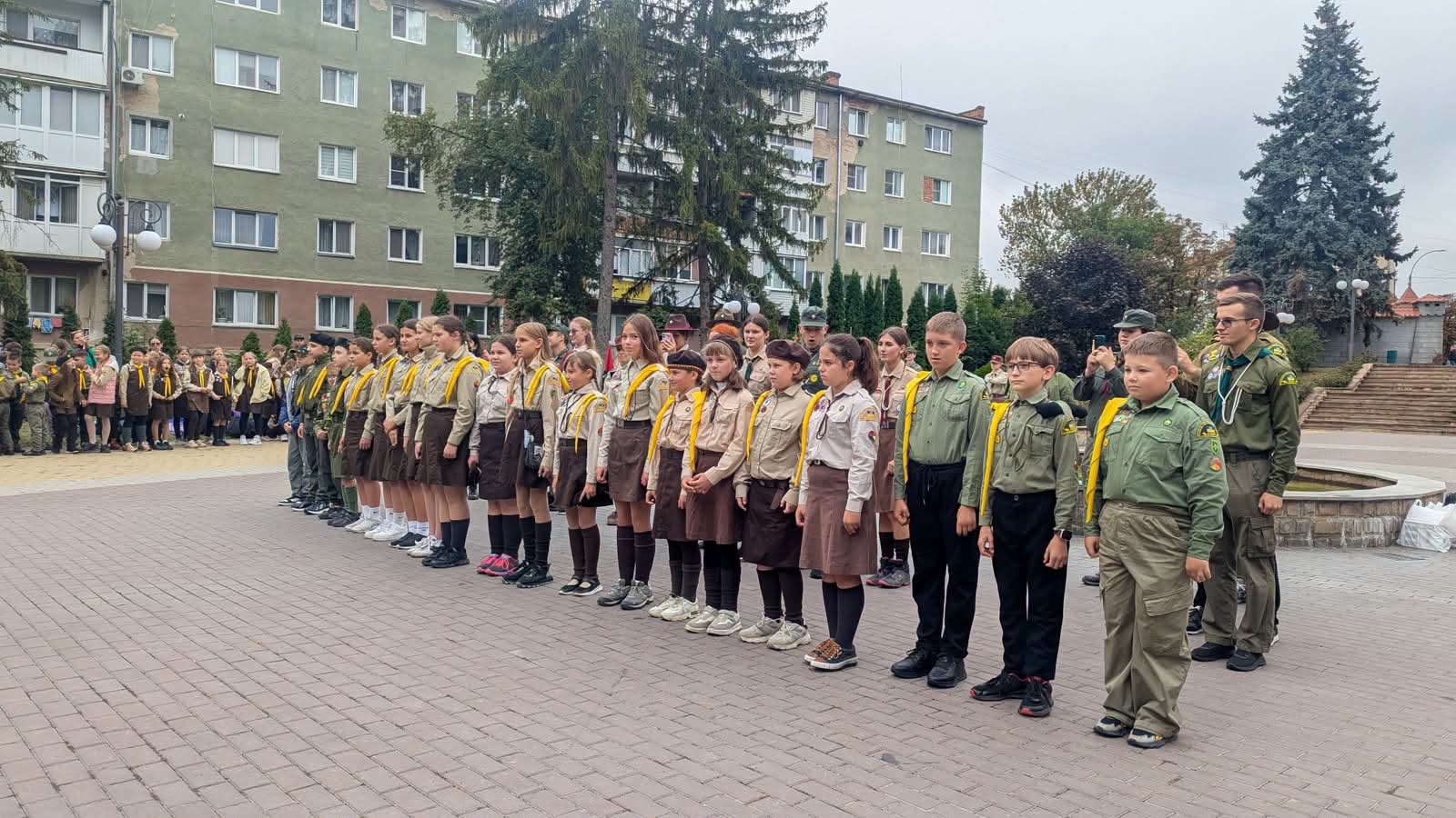Група юних пластунів і пластунок стоїть у рівному ряду на бруківці. Діти одягнені в пластову форму з жовтими хустками. Позаду багатоквартирні будинки, дерева. Дехто з дорослих стоїть збоку.