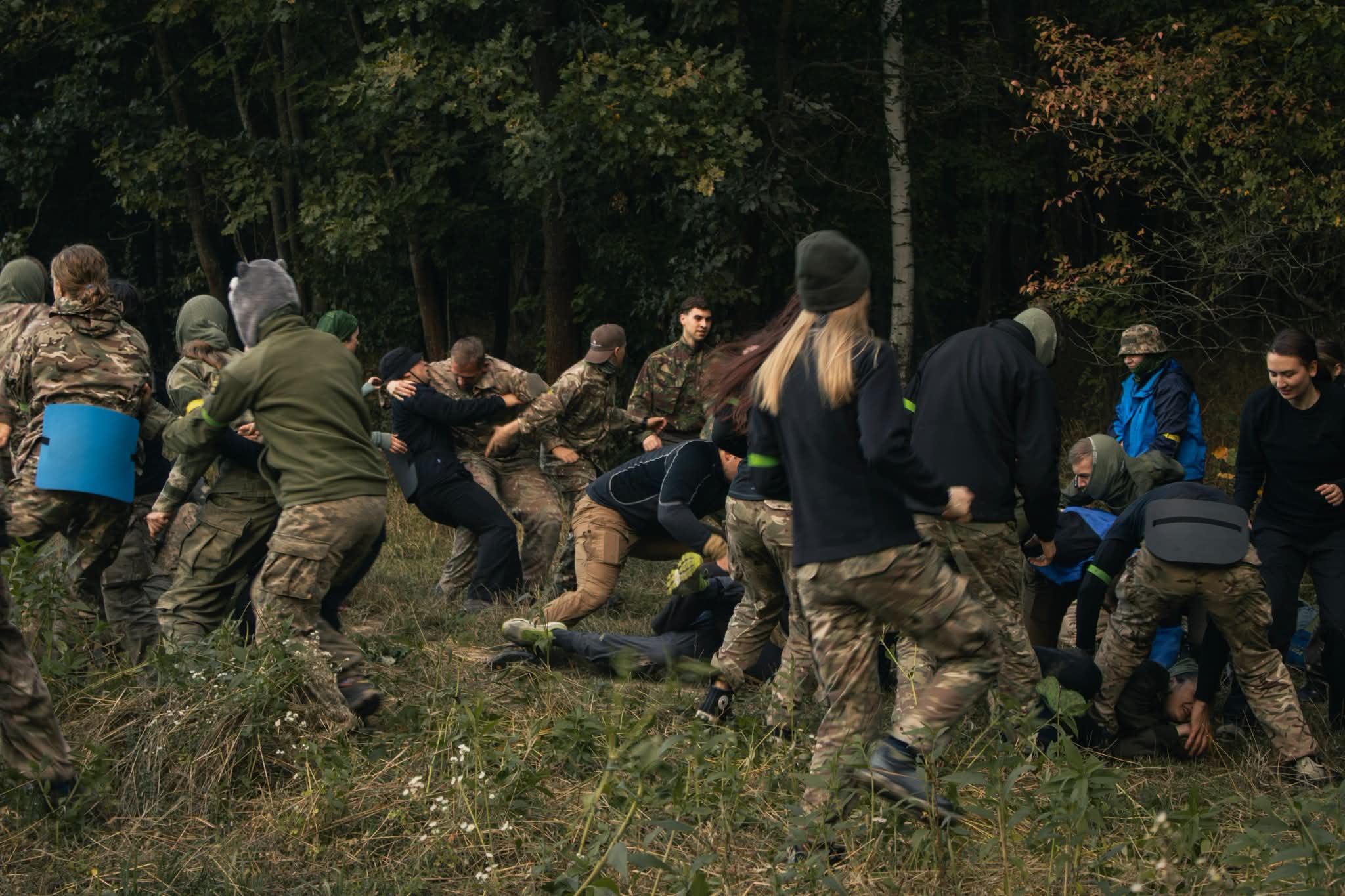 Учасники в камуфляжі та спортивному одязі активно взаємодіють під час гри в лісі. Частина з них тримає захисні щити.