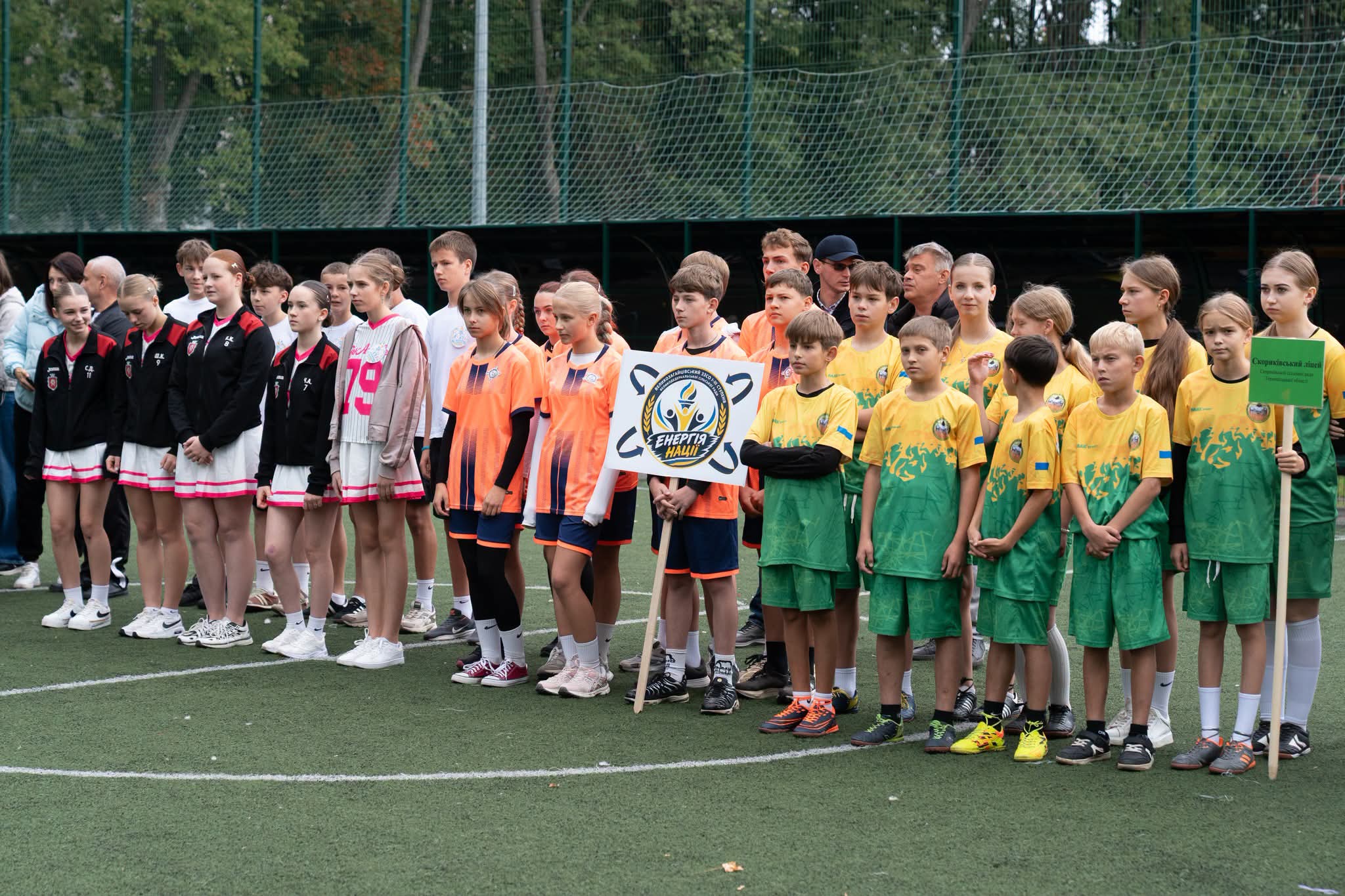 Група дітей у різнокольоровій спортивній формі вишикувалася на футбольному полі. Деякі тримають таблички з написом &laquo;Енергія нації&raquo; та назвою школи.