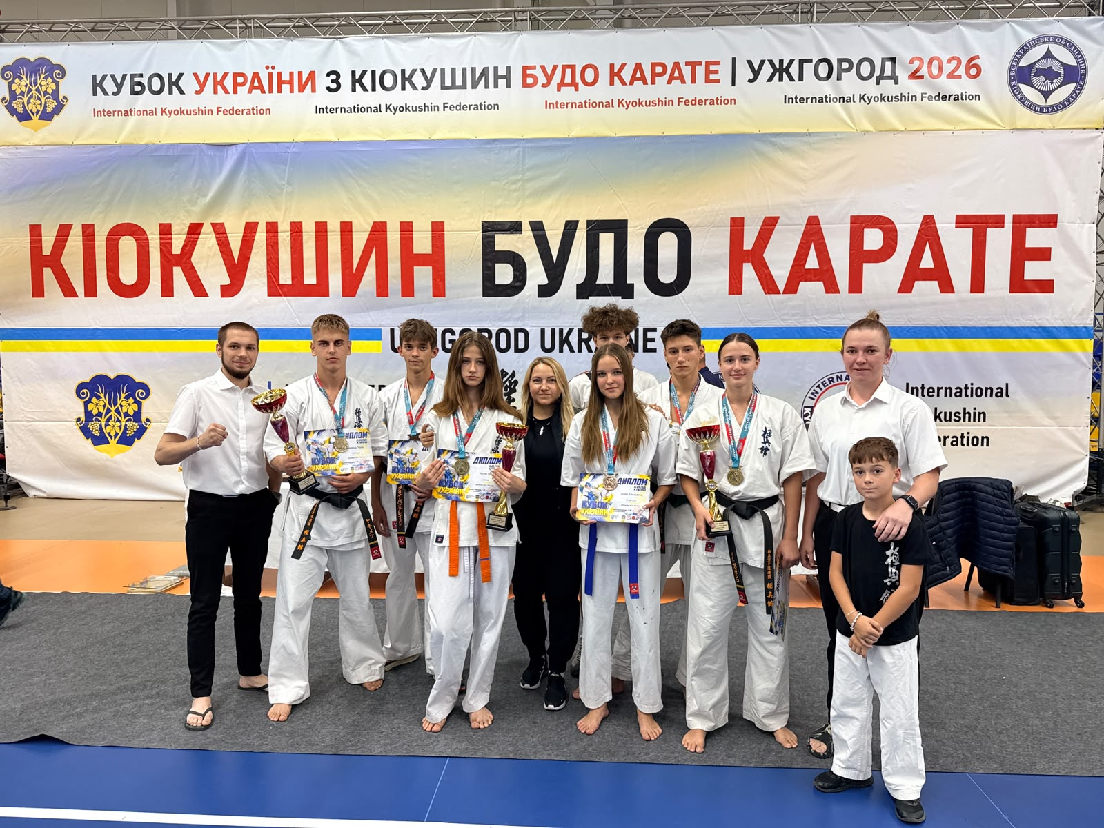 Групове фото учасників і тренерів після змагань на синьому татамі в спортивному залі. Позаду великий банер з написом «Кубок України з кіокушин будо карате». У центрі стоять спортсмени в білих кімоно з поясами різних кольорів (чорні, помаранчеві, сині), які тримають кубки, медалі та дипломи.