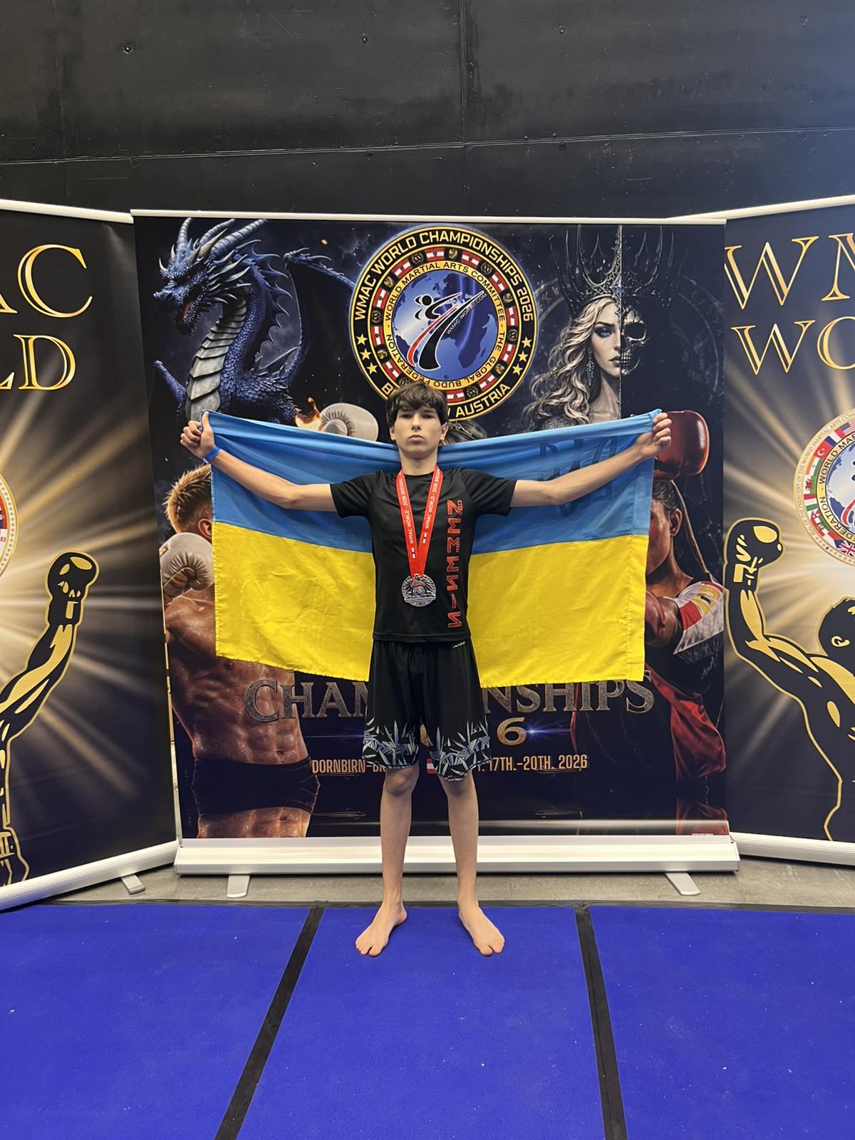 Хлопець у чорній спортивній формі стоїть босоніж і тримає розгорнутий український прапор. На шиї в нього висить медаль Чемпіонату світу.