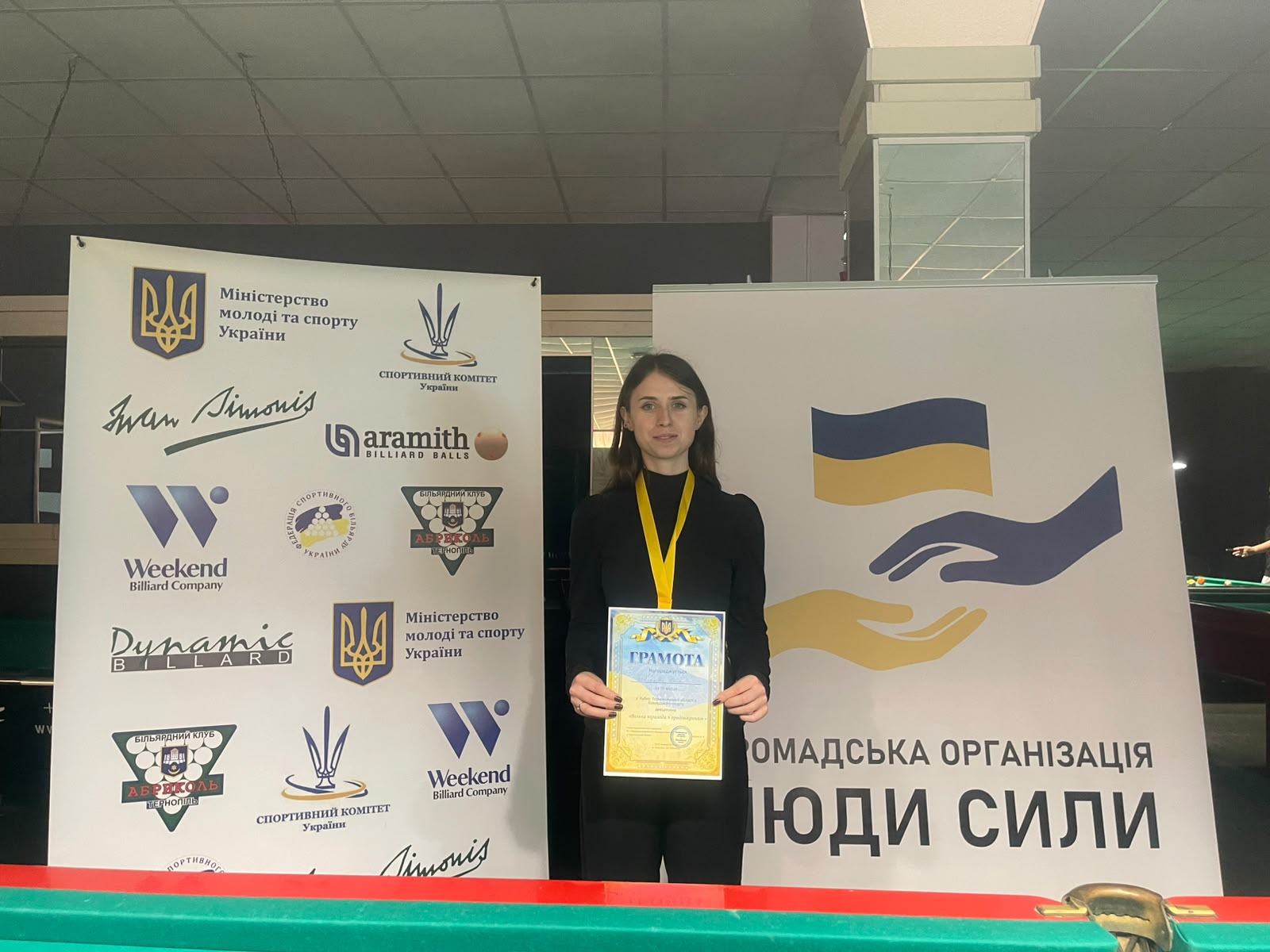 Юлія Малахова (Тернопіль), бронзова призерка турніру, тримає грамоту. На ній медаль на жовтій стрічці, позаду &mdash; банери клубу та Міністерства молоді та спорту.