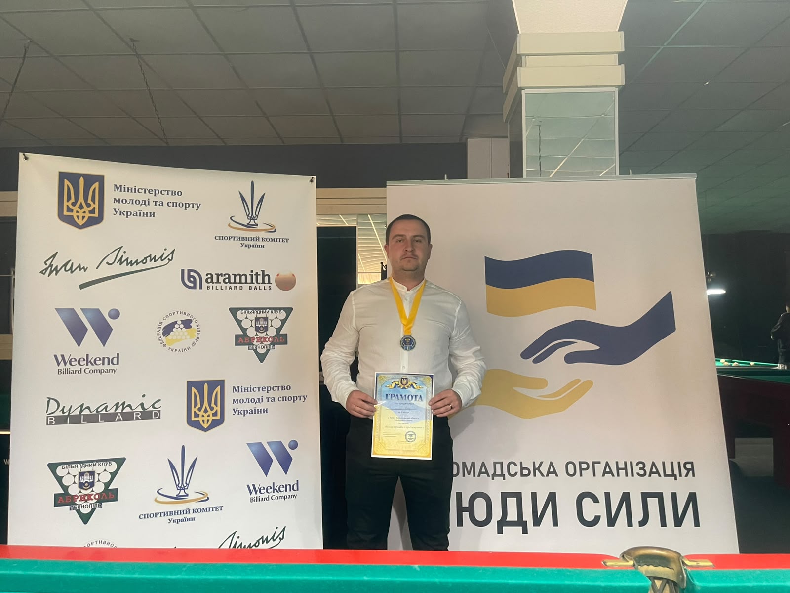 Владислав Мусієнко (Київ), срібний призер турніру, усміхається з кубком і грамотою в руках. На ньому медаль на жовтій стрічці, на фоні &mdash; рекламні банери спонсорів.