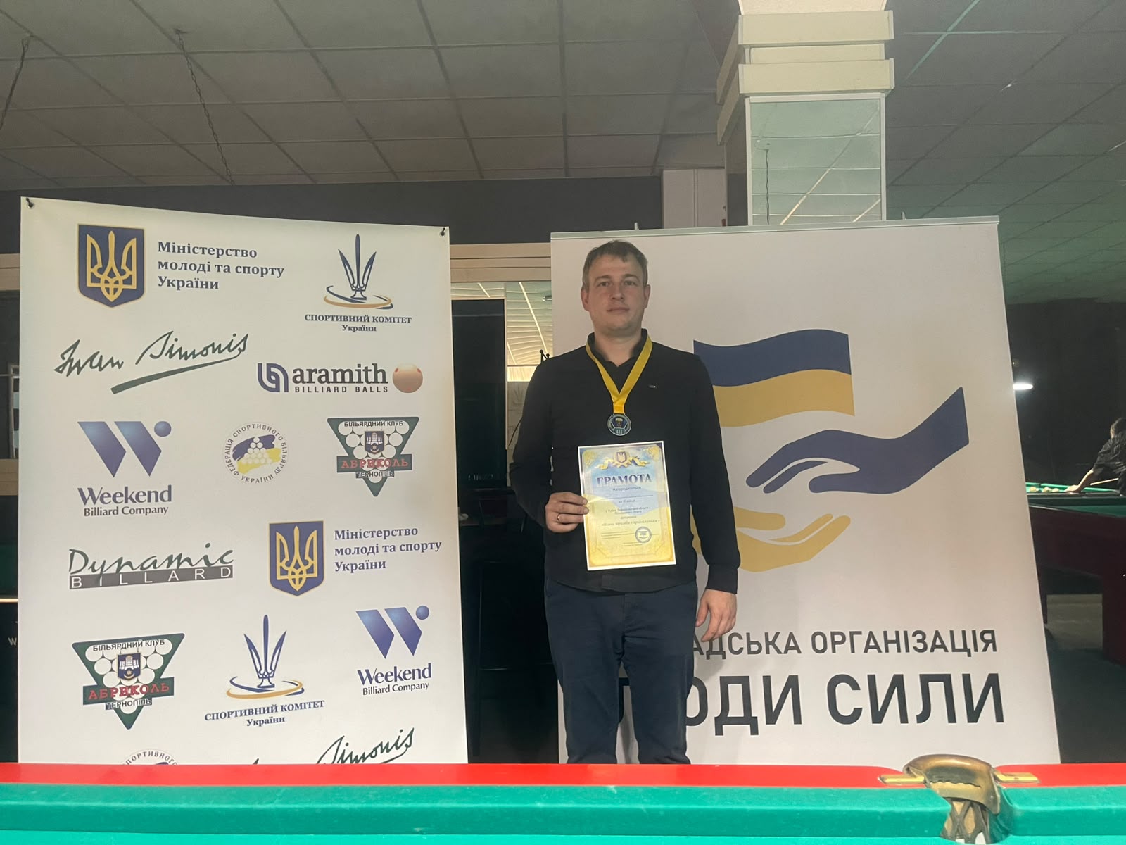 Олександр Сохарєв (Тернопіль), бронзовий призер змагань, тримає грамоту з медаллю на шиї. Він стоїть перед банерами спортивних організацій.