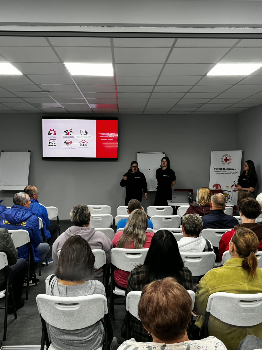 Дві жінки в чорному одязі стоять біля екрана з іконками послуг Громадського центру Червоного Хреста і проводять презентацію. Учасники сидять спиною до камери і слухають