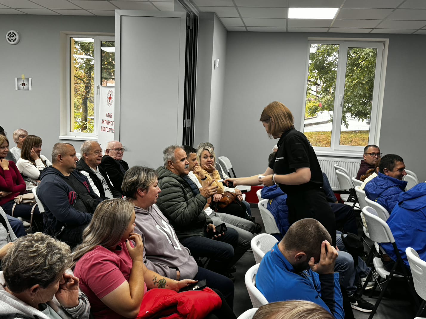 Жінка в чорному одязі нахиляється до учасників у рядах і щось показує. Слухачі уважно дивляться на неї